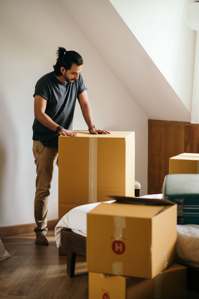 Ethnic man packing belongings in cardboard boxes while standing in modern apartment with white wall and looking down while preparing for relocation